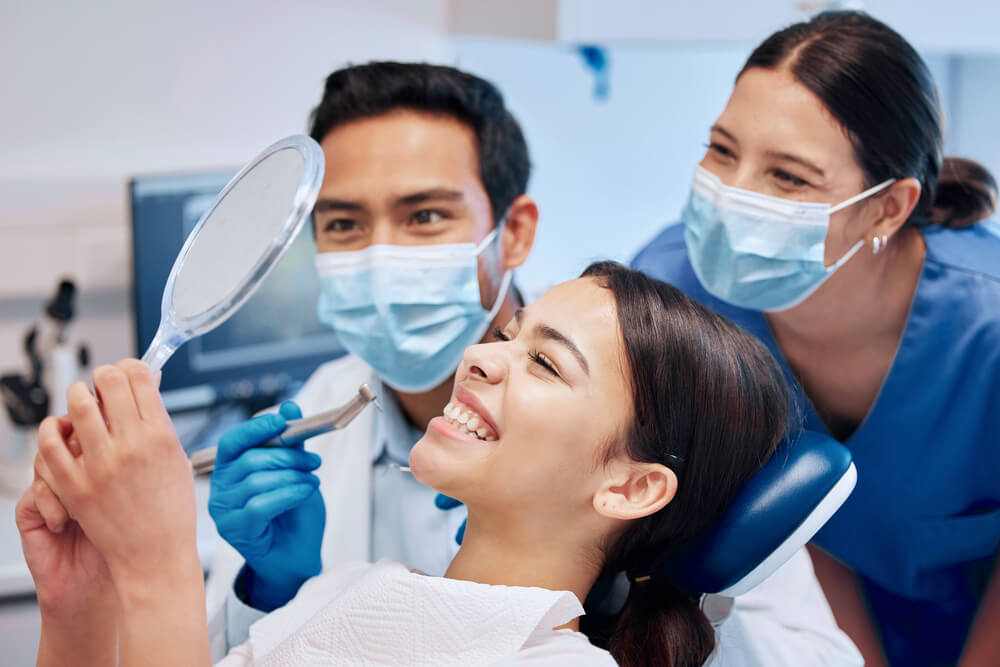 A woman patient gazes at the mirror while the dentist and assistant eagerly anticipate her smile.
